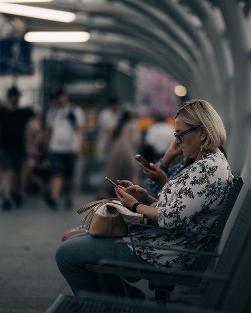 Free Blonde woman sitting indoors, using a smartphone at a train station. Busy background. Stock Photo
