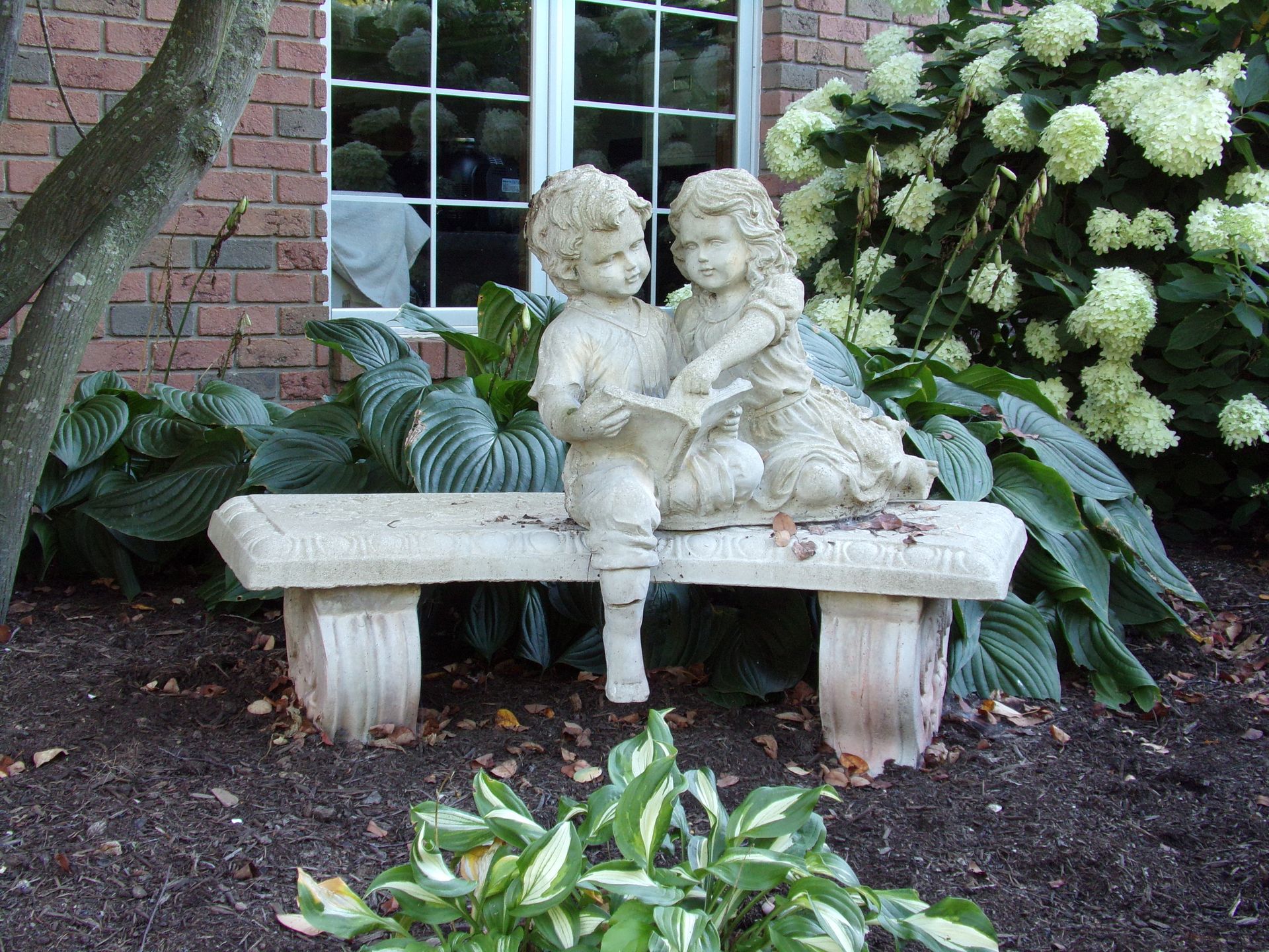 Beautiful stone statue of children on a bench surrounded by lush garden foliage.