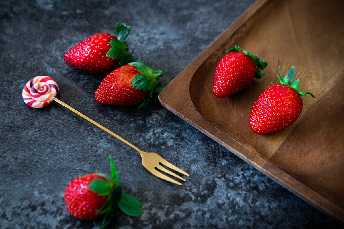 Close-up of fresh strawberries beside a candy on a small gold fork, placed next to a wooden plate.