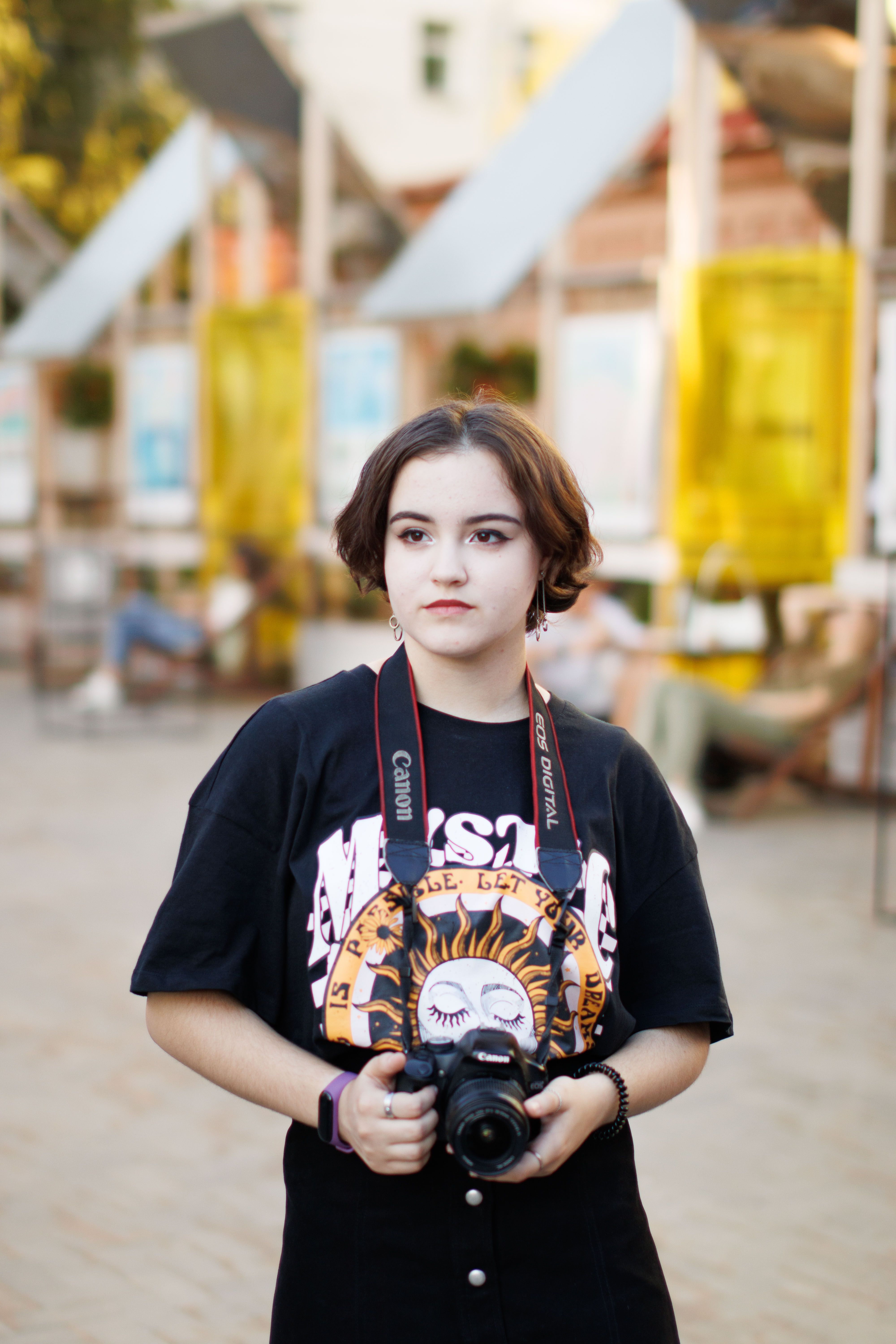 Young woman holding a Canon camera outdoors, dressed casually in a city environment.