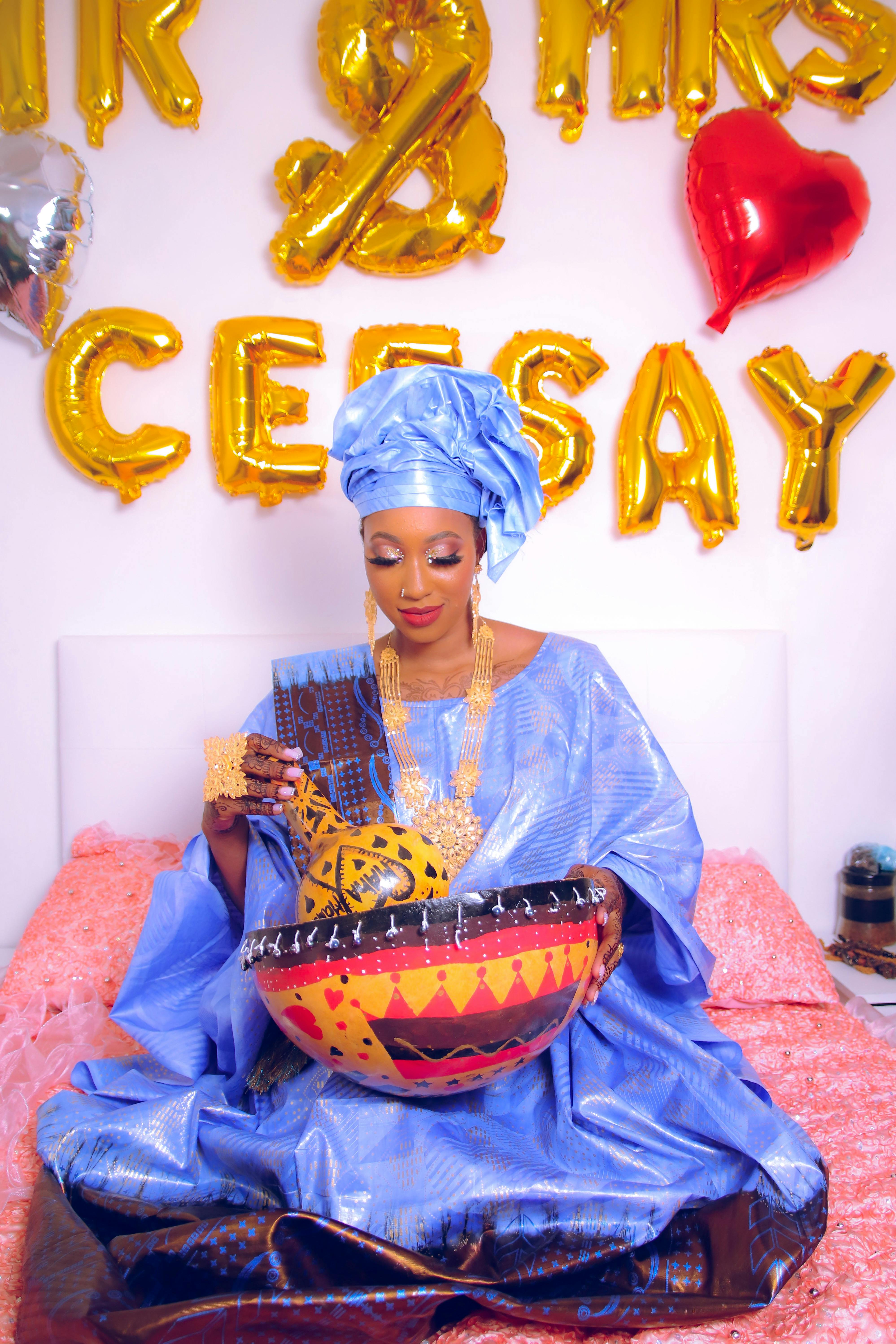 Graceful woman in traditional blue clothing, holding an intricately designed bowl, seated against celebratory balloons.