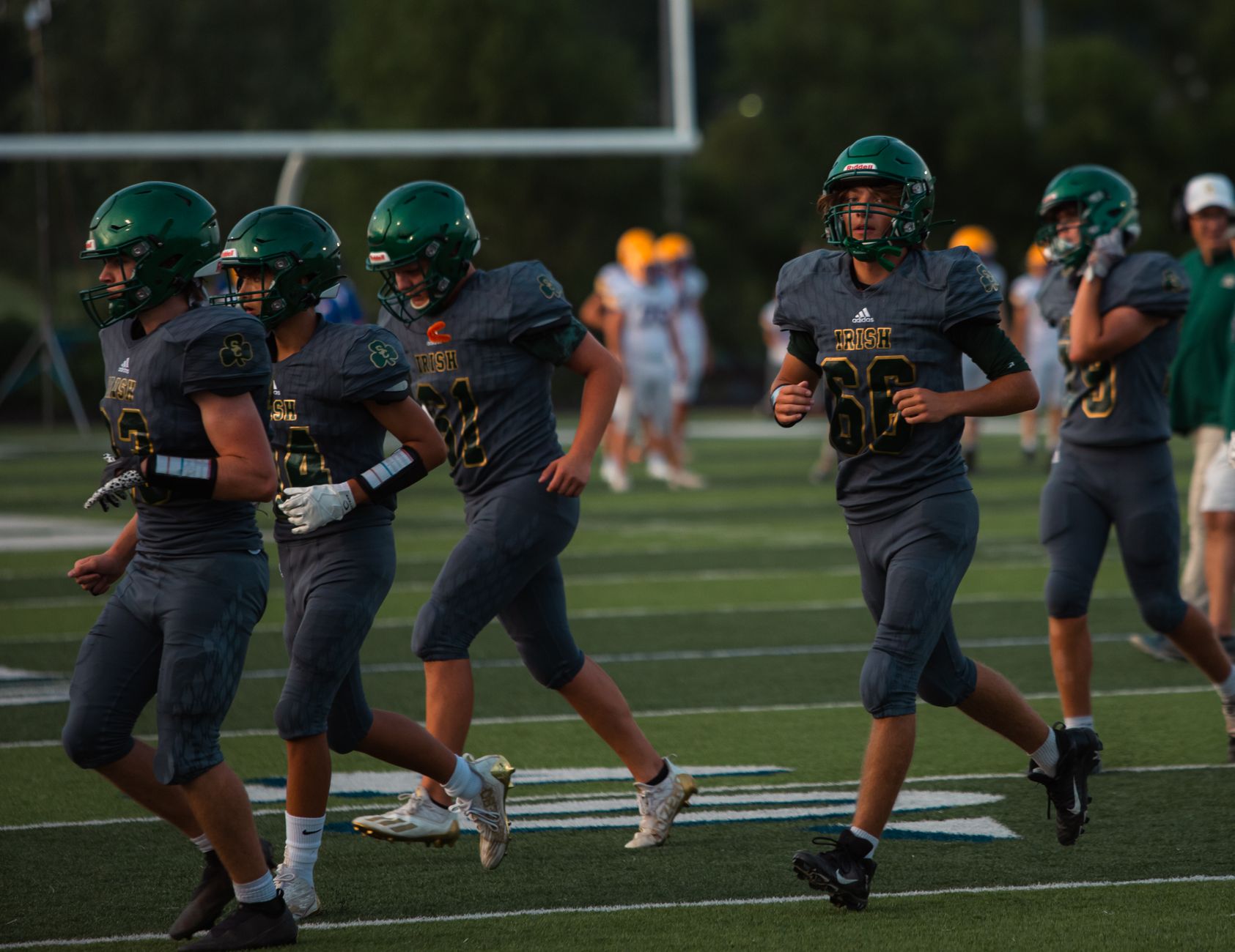 High school football team running on field during sunset, wearing green uniforms and helmets.