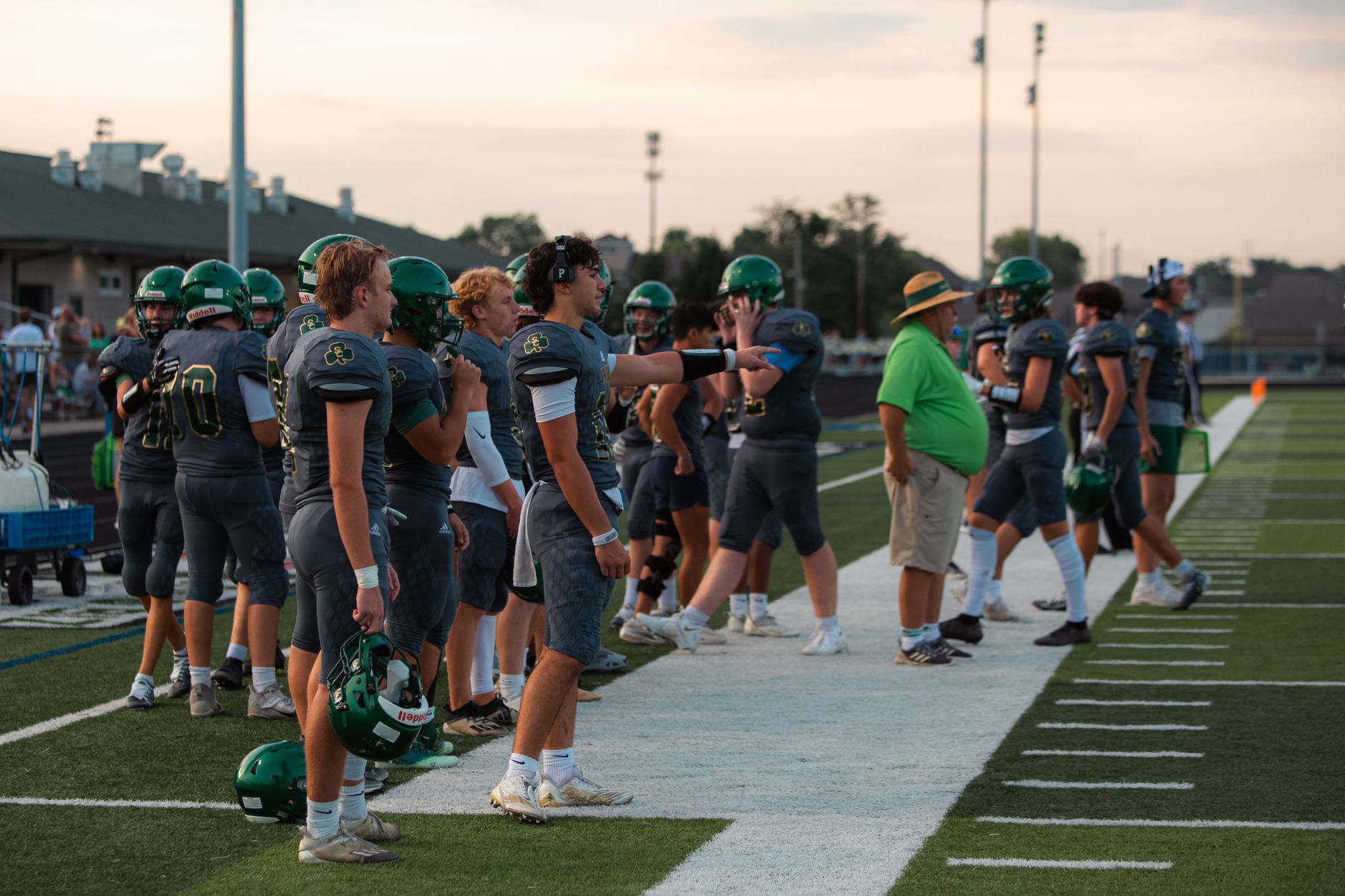 High school football players on the sidelines during a game preparation at sunset.