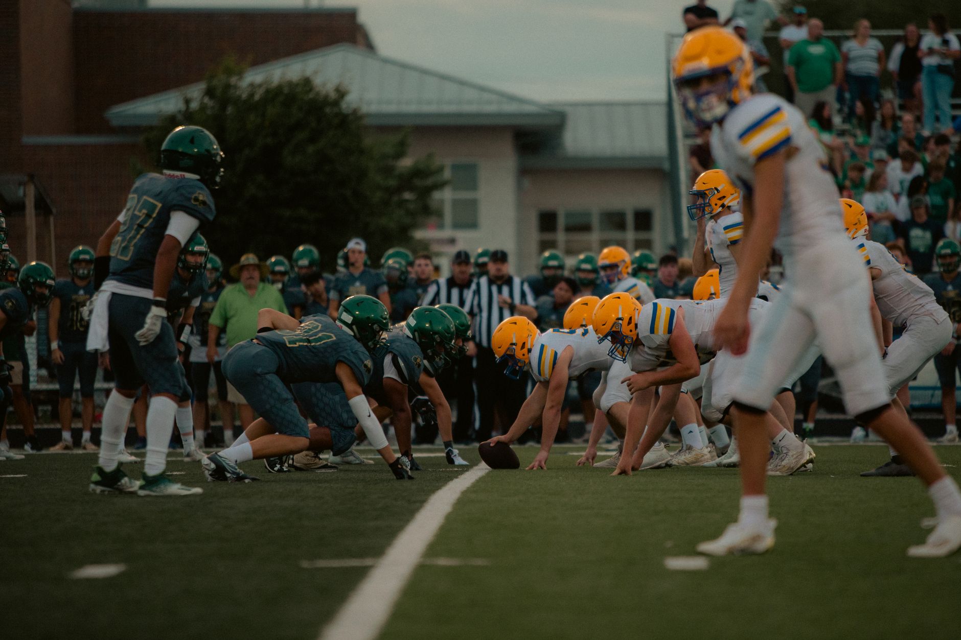 High school football teams face off with players in action under evening lights.