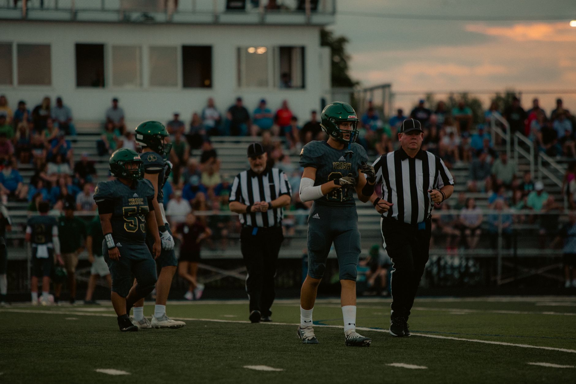 high school football officials discussing player conduct beside a crowded sideline during practice
