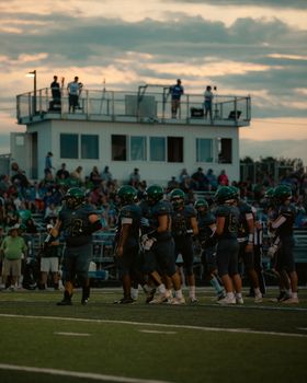 A high school football team on the field with a crowd in the background during sunset.