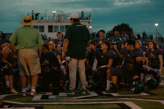 Football team huddle with coaches during an evening game at an outdoor stadium.