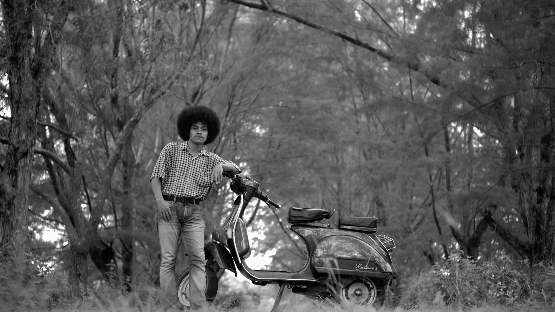 Black-and-white photo of a man with an afro hairstyle posing with a classic Vespa scooter outdoors.