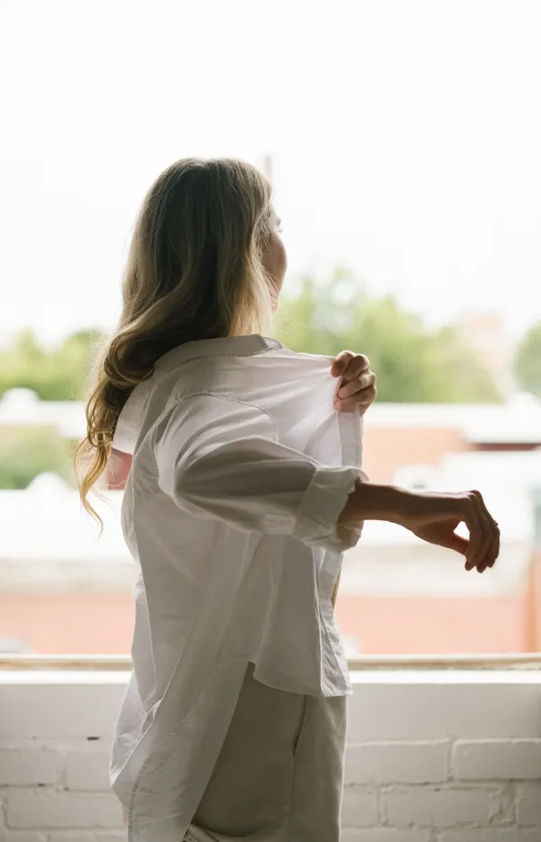 Woman putting on a loose white shirt by a window