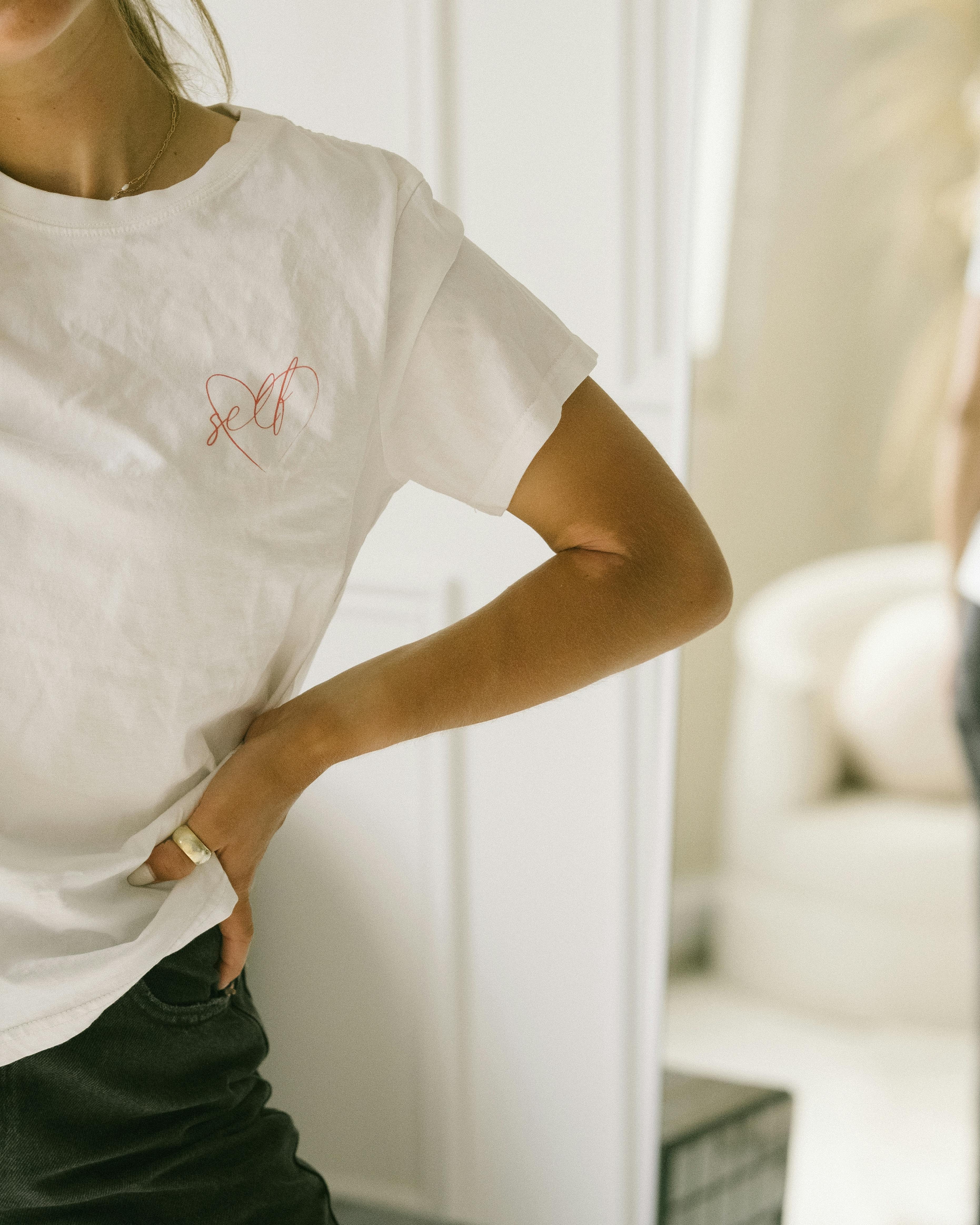 Close-up of a woman in a white t-shirt with hand on hip in a cozy indoor setting.
