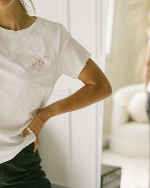 Close-up of a woman in a white t-shirt with hand on hip in a cozy indoor setting.
