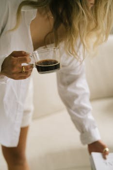 A woman holding a glass cup of coffee, dressed in a casual white shirt, creating a cozy morning vibe.