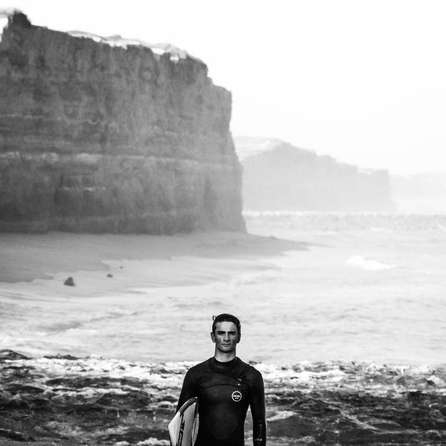 Monochrome photo of a surfer with a board on a rugged beach cliff landscape.