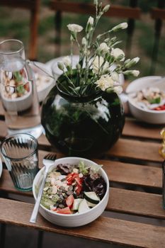 A fresh salad in a white bowl placed on a wooden table with a vase of white flowers and glassware.