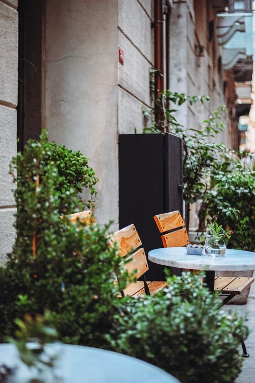 Free Cozy outdoor cafe scene with wooden chairs, tables, and lush greenery in an urban setting. Stock Photo