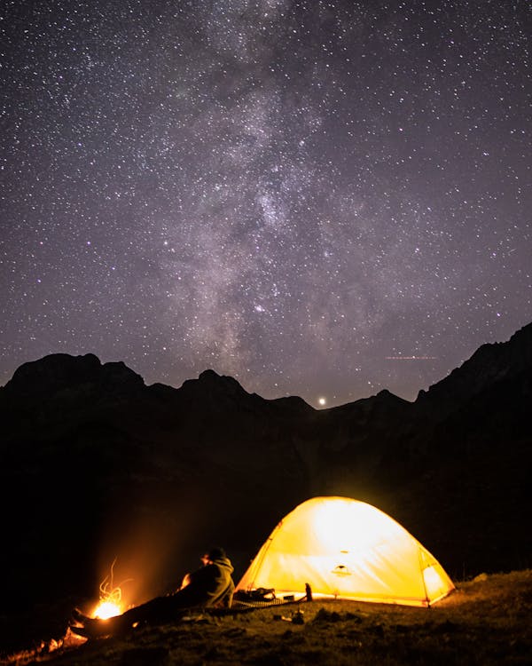 Man Lying Next to a Bonfire and a Tent in Hills Under a Starry Sky