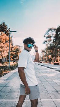 A fashionable man wearing sunglasses poses confidently in a sunny outdoor setting, exuding style and attitude.