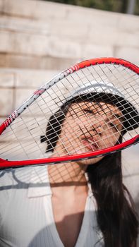 A young woman poses with a tennis racket outdoors on a sunny day, casting interesting shadows.