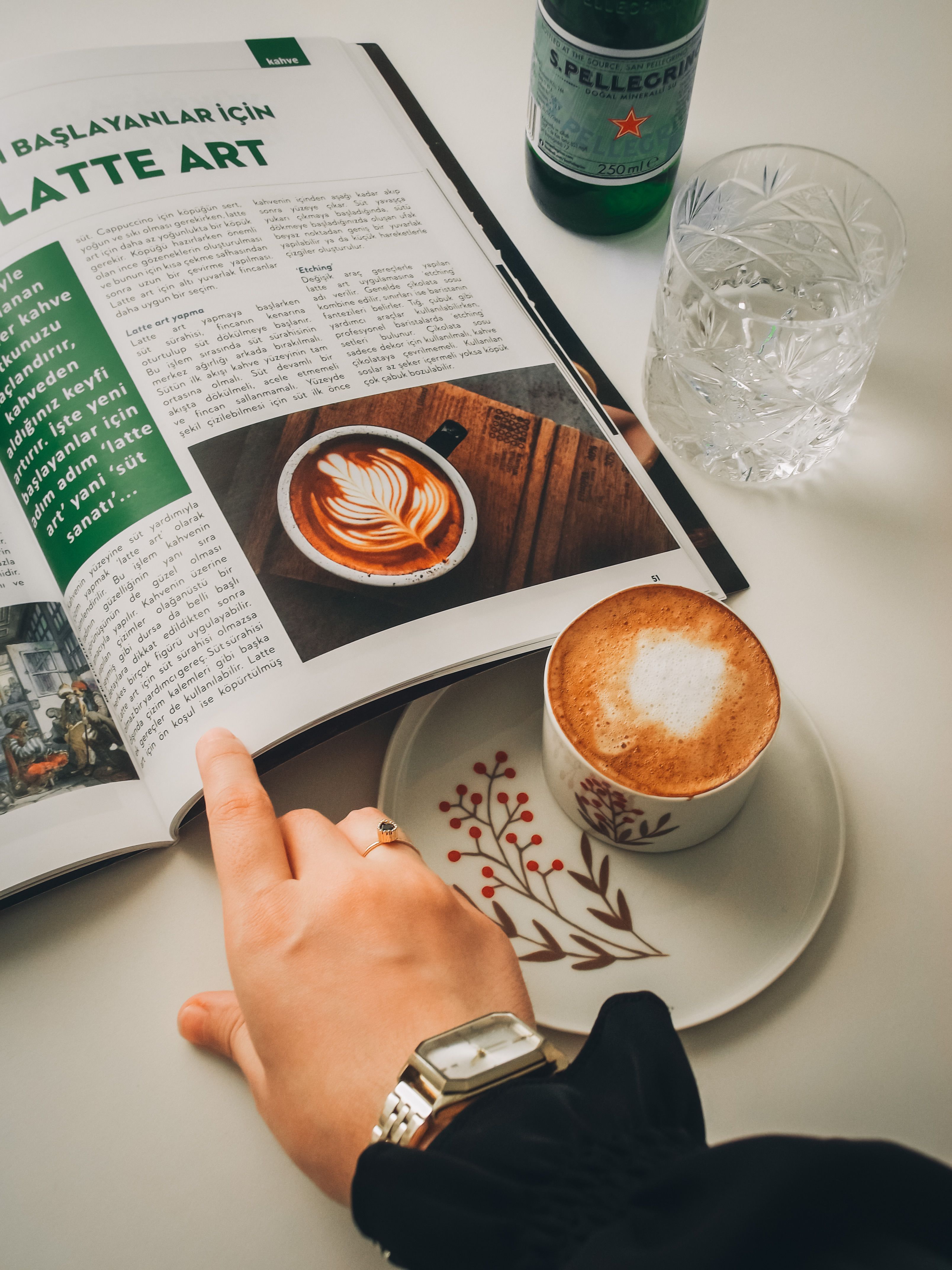 Hand holding a latte art magazine open beside a cappuccino with creative foam design on a white ceramic cup.