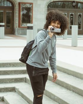 Smiling young man with afro hair holding a camcorder on steps outdoors.