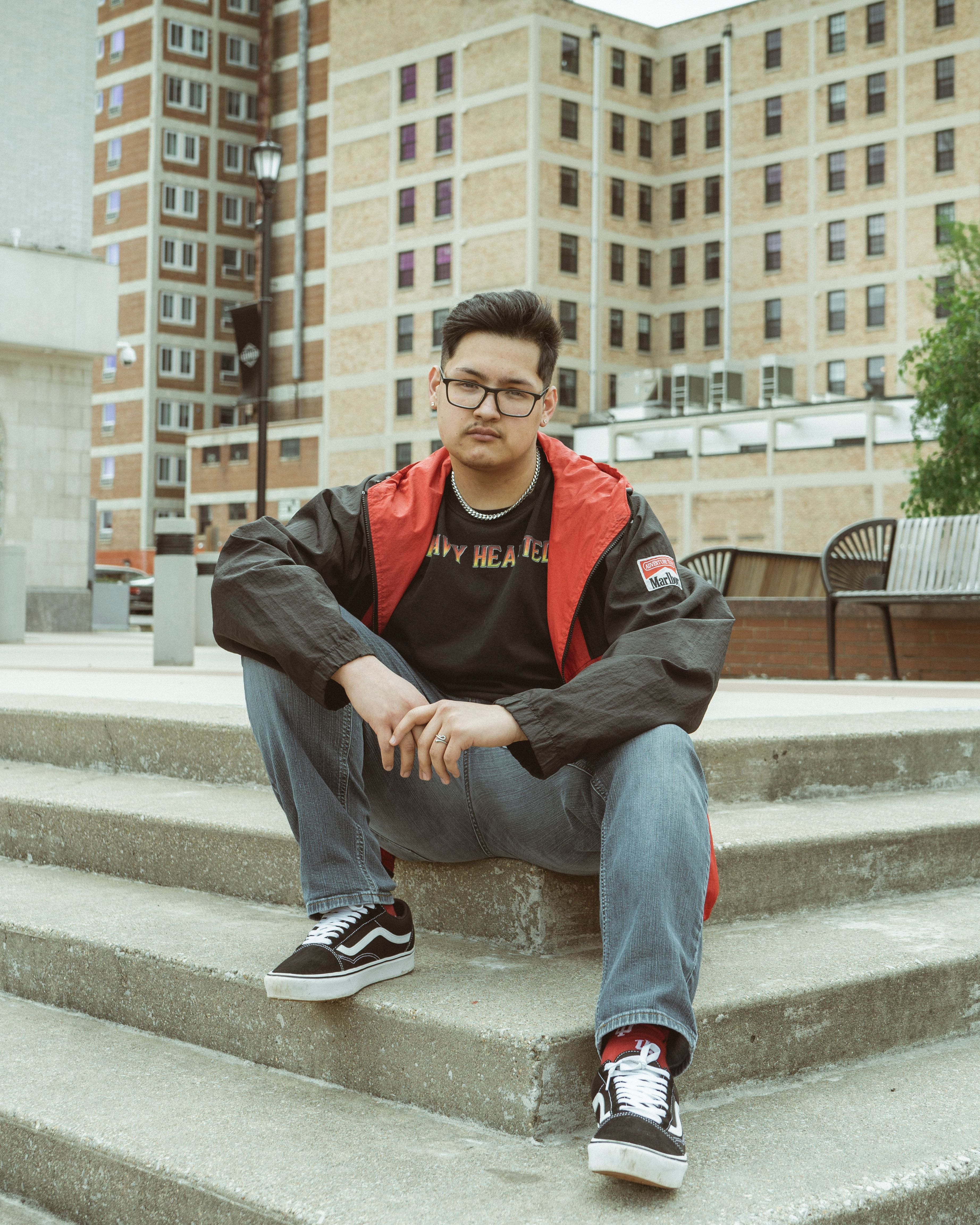 Asian man in a black jacket and denim posing on outdoor concrete steps, trendy city fashion.