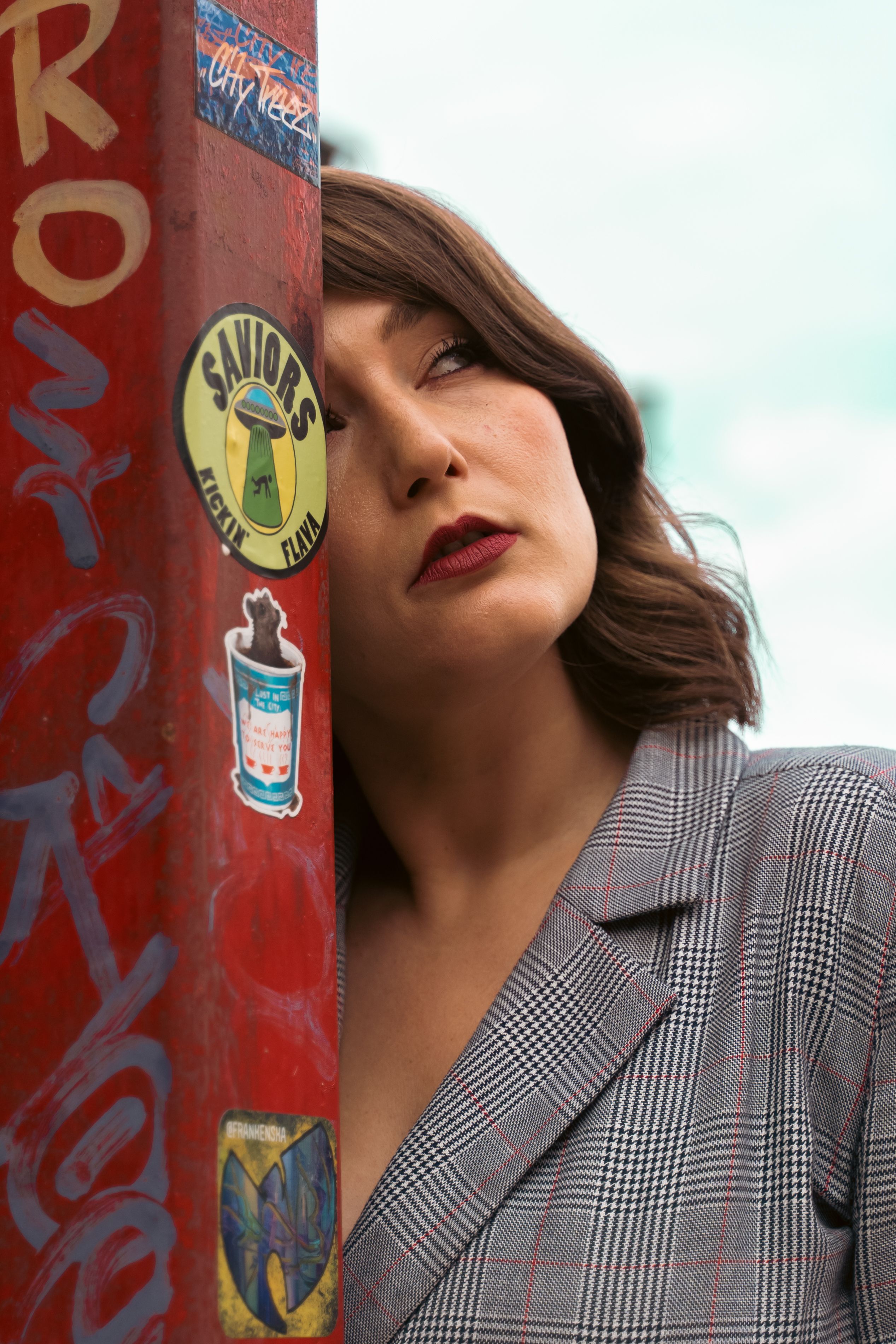 A stylish woman leans near a graffiti-covered pole, showcasing urban fashion with a plaid blazer and red lipstick.