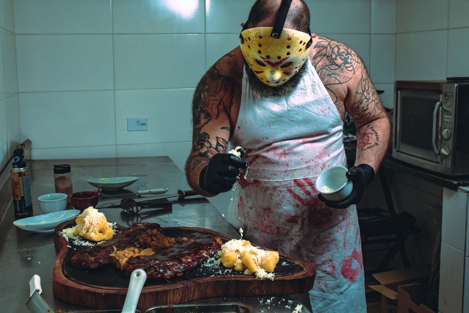 Como Estimular a Criatividade na Cozinha com Receitas Fáceis 2 A tattooed man in a horror mask garnishes food in a kitchen. Creepy culinary art.