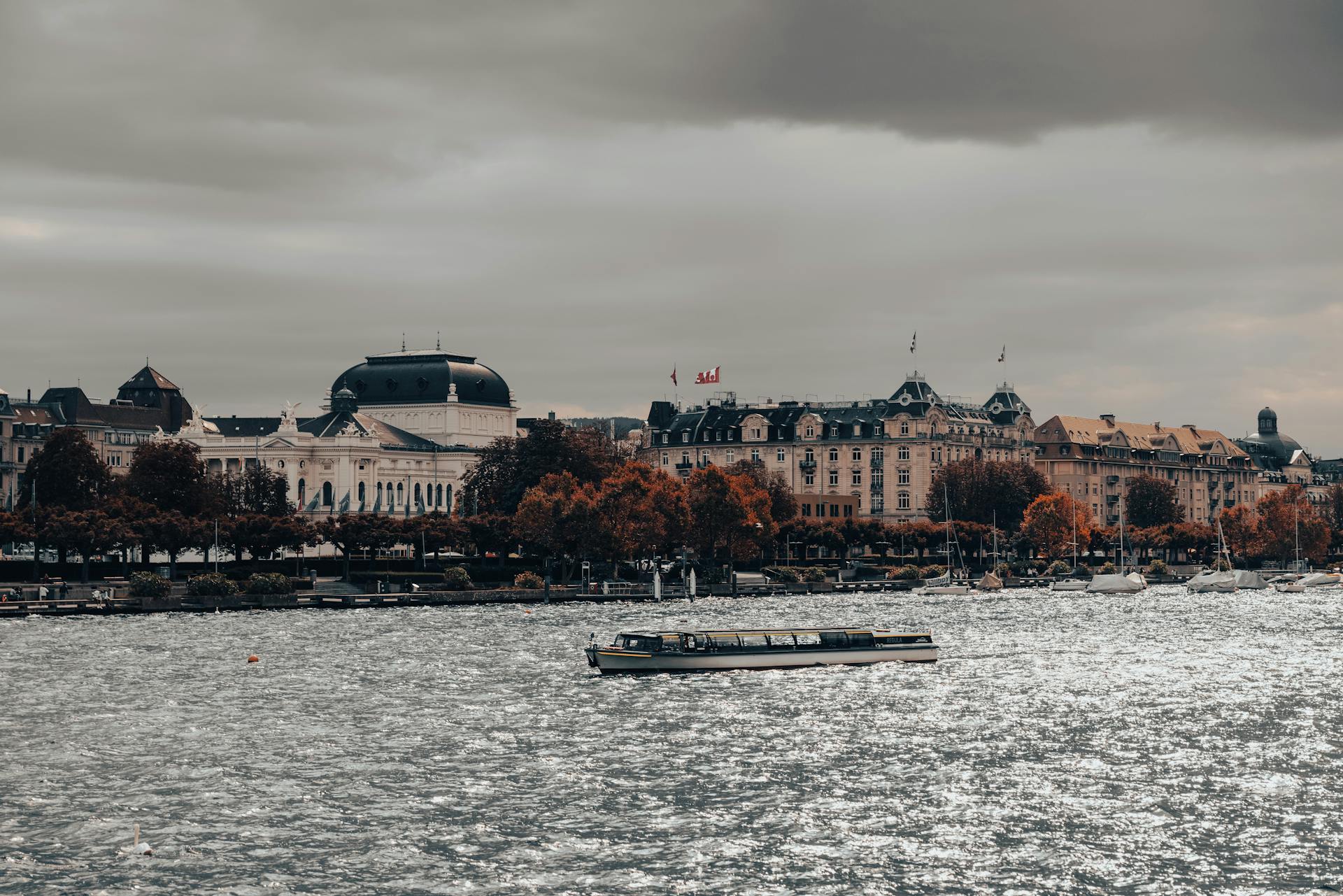 A scenic view capturing buildings and a boat along Lake Zurich under a cloudy sky.