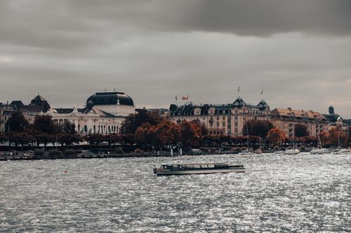 Free A scenic view capturing buildings and a boat along Lake Zurich under a cloudy sky. Stock Photo
