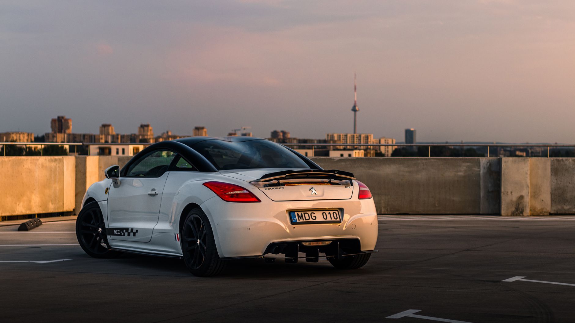 A sleek white Peugeot parked on a rooftop with a cityscape during sunset.