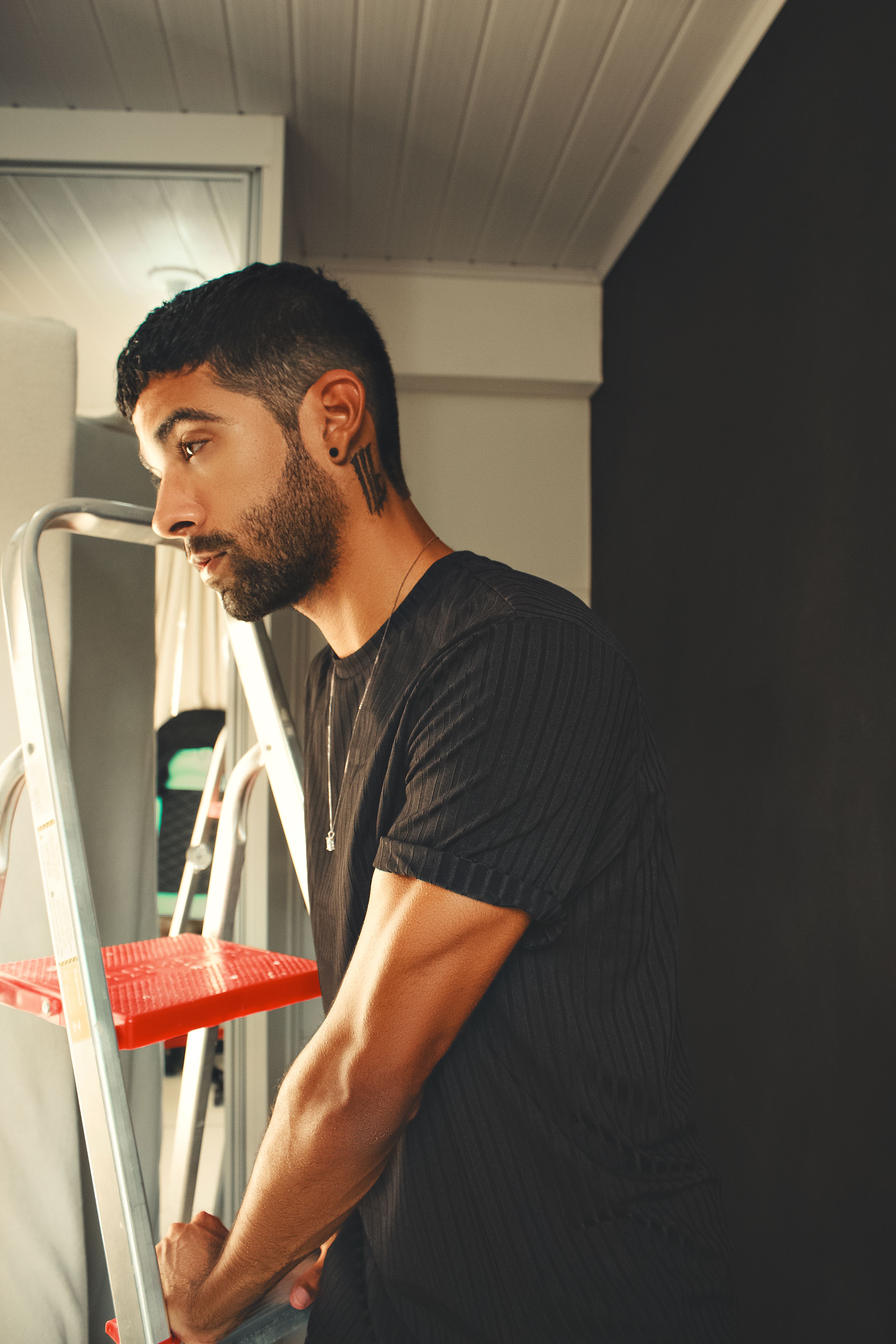Bearded man wearing black shirt standing next to a ladder indoors, facing mirror.