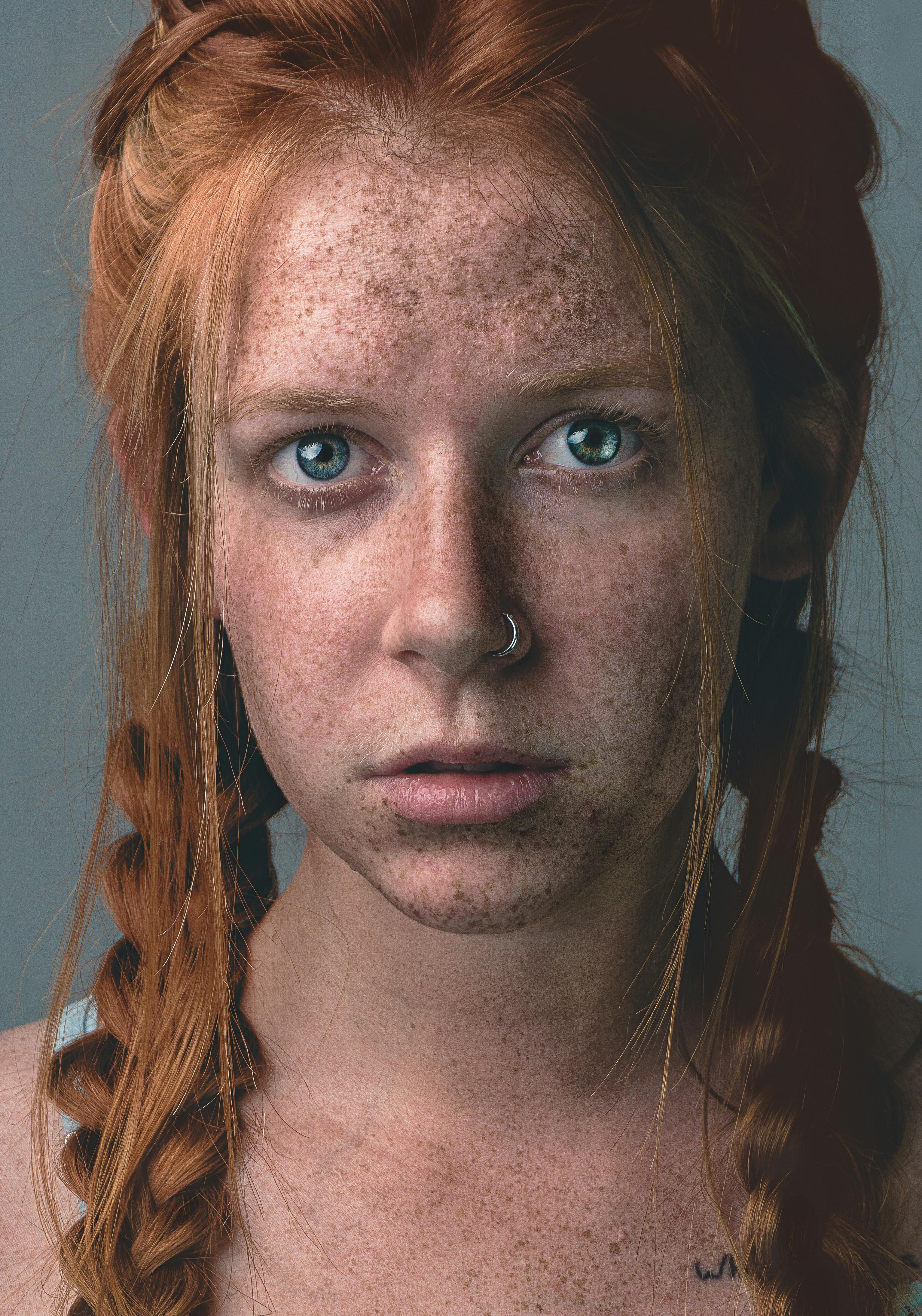 Close-up of a young woman with freckles, braided red hair, and a nose piercing.