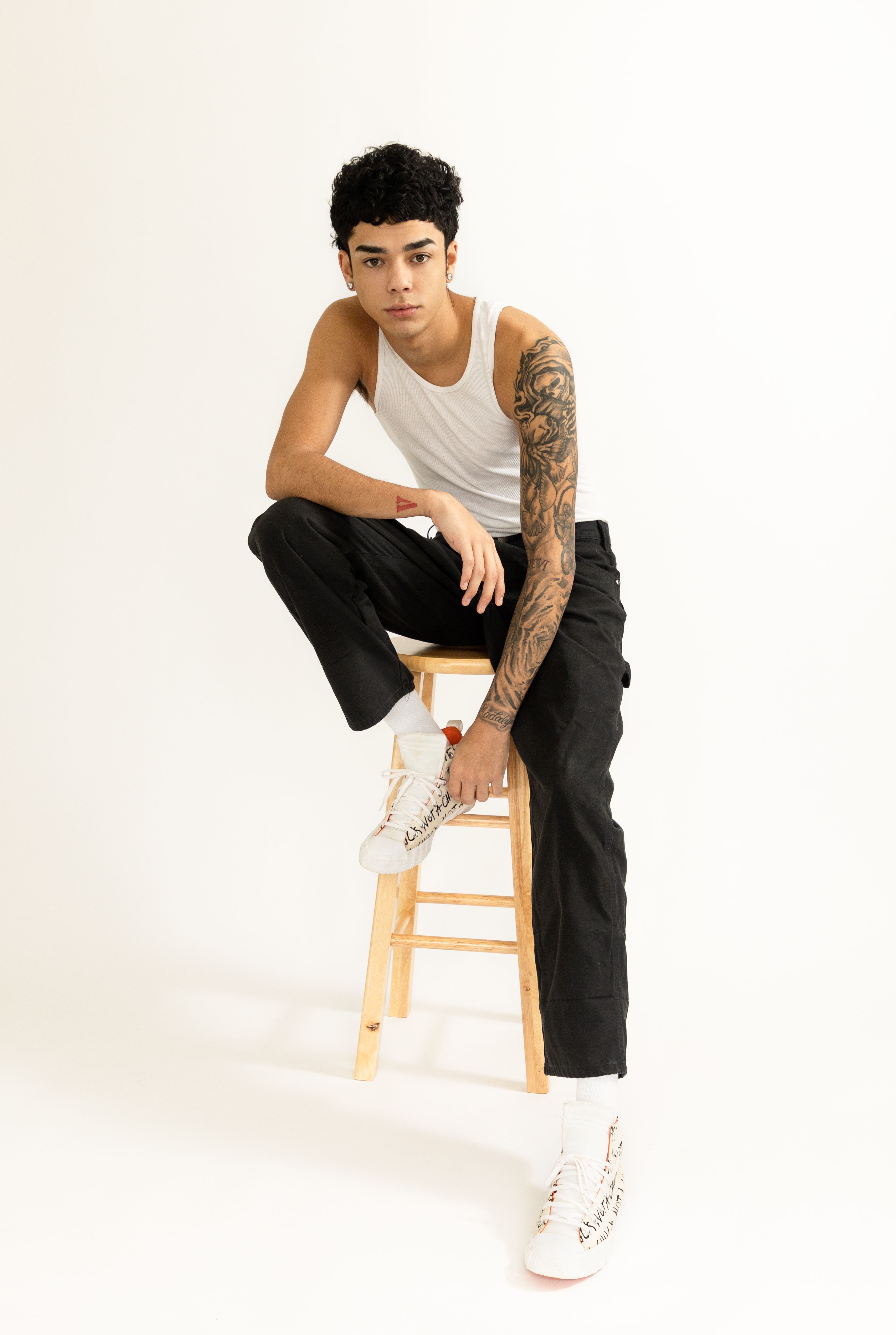 Studio shot of a young man with a striking arm tattoo sitting on a stool in a minimalist setting.