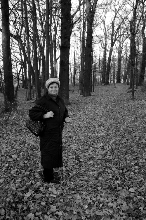 Grayscale Photo of an Elderly Woman Standing on Leaf Covered Ground