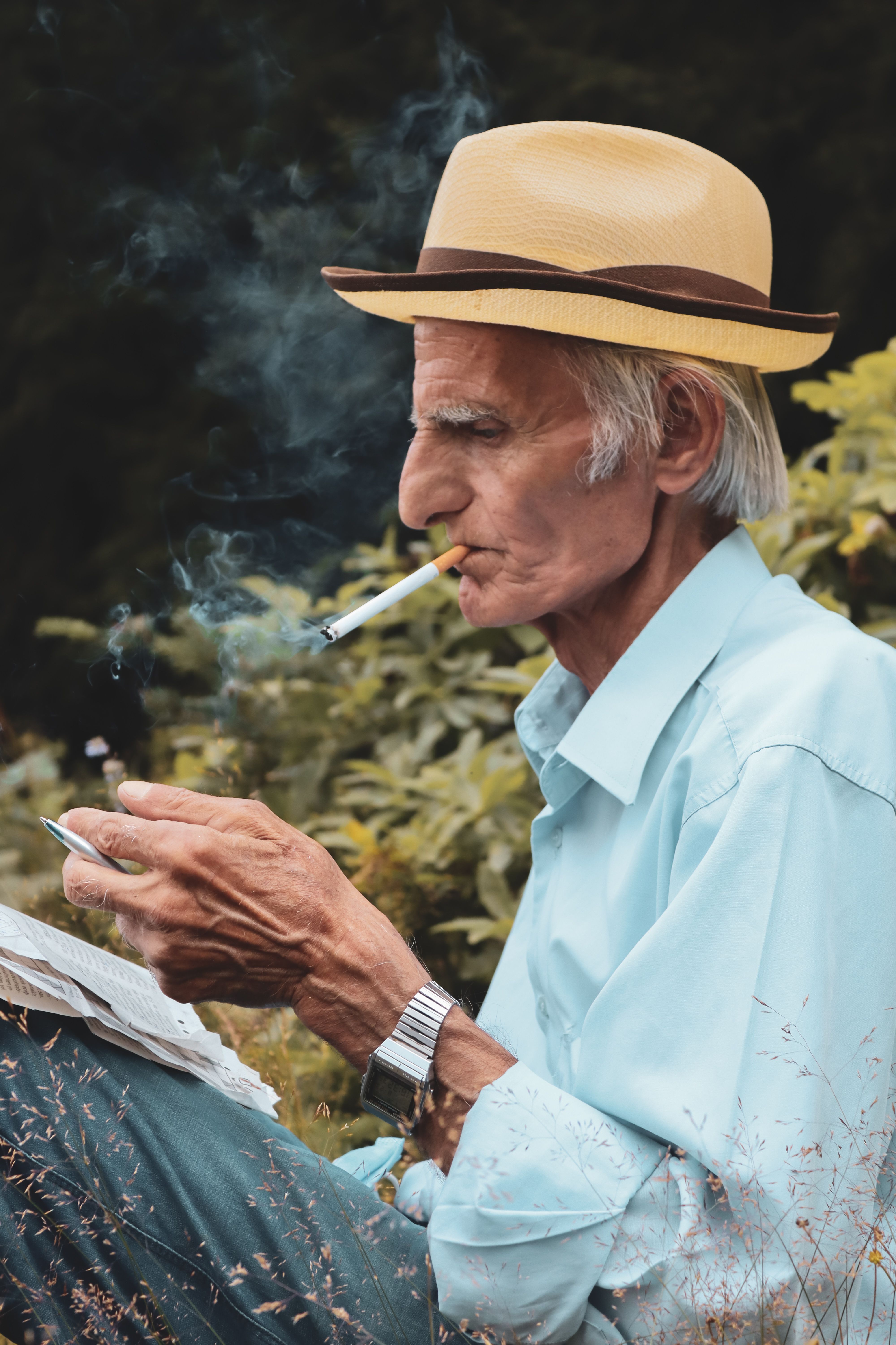 Senior man in a hat smokes a cigarette outdoors, enjoying a relaxing moment.