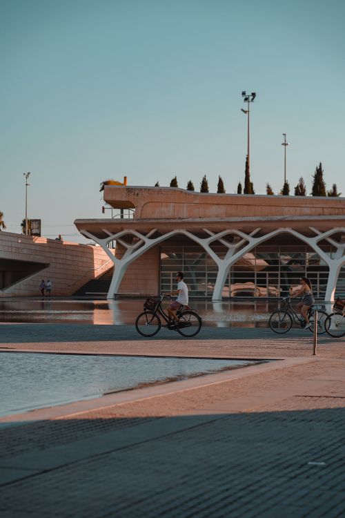 Free A serene scene featuring people biking beside a modern architectural structure on a sunny day. Stock Photo