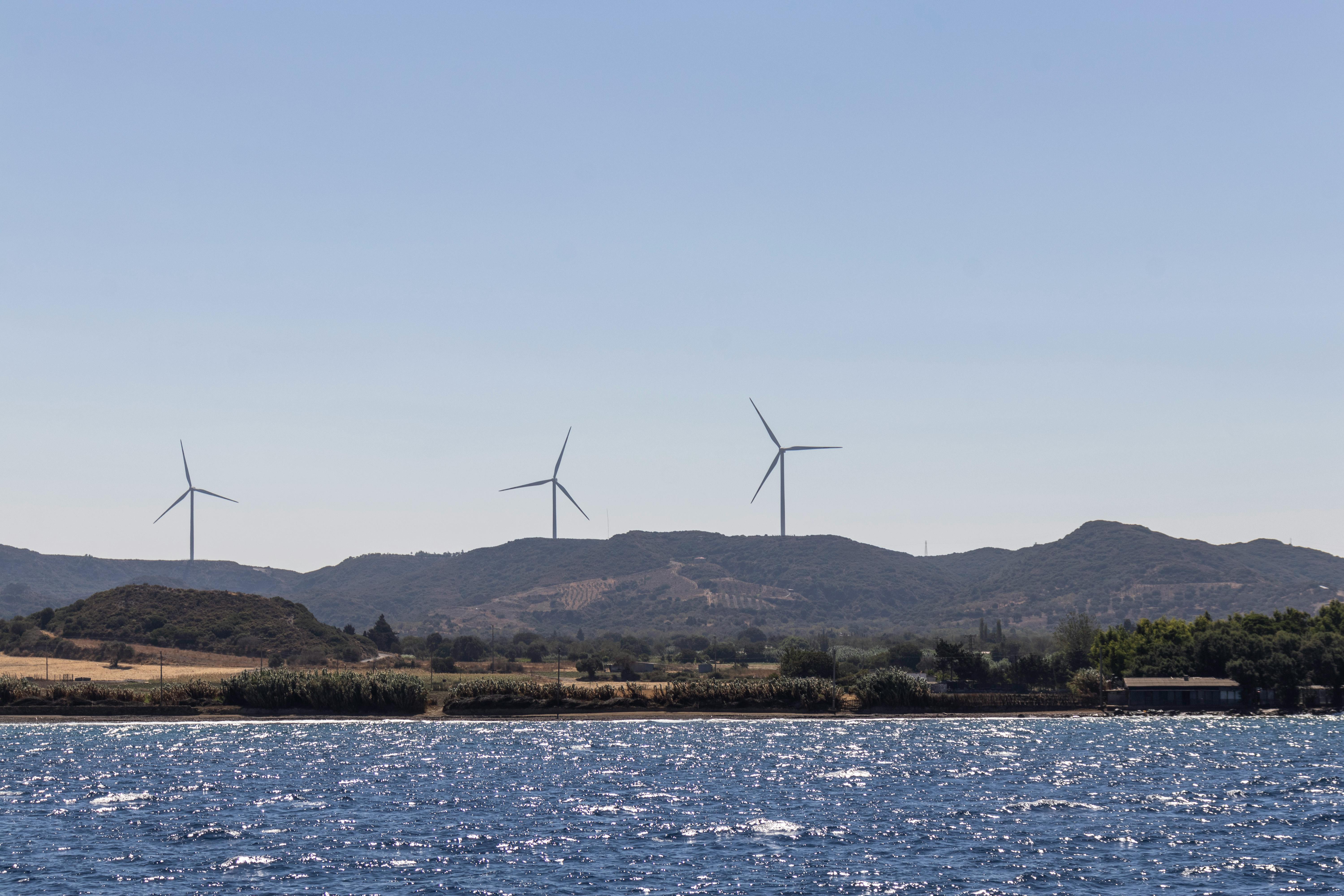 Wind turbines on a hillside with a sparkling sea under a clear blue sky, showcasing renewable energy.