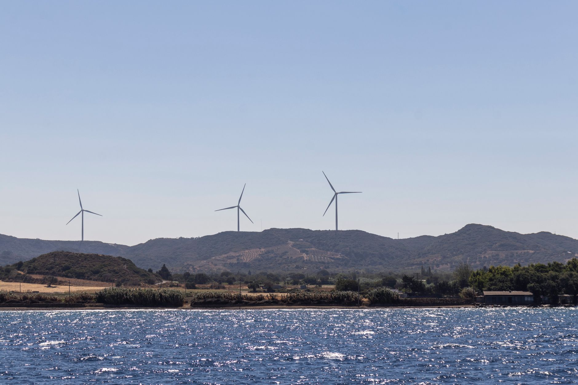 Wind turbines on a hillside with a sparkling sea under a clear blue sky, showcasing renewable energy.