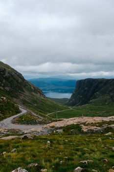 Curved asphalt road through lush green mountains leading to a stunning lake view under cloudy skies.