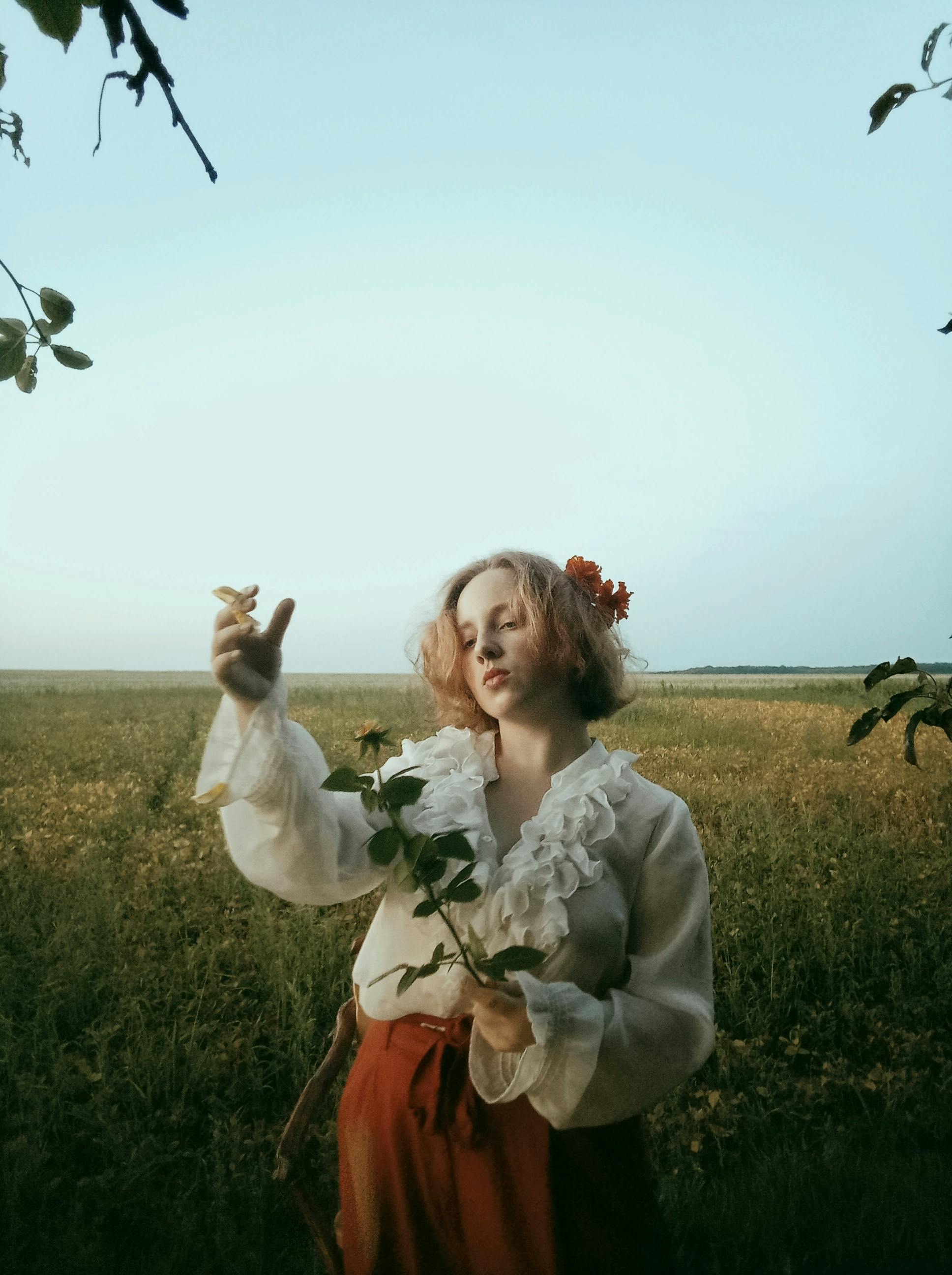 A young woman in vintage attire holds a flower in a serene, rustic field, evoking a timeless, peaceful mood.