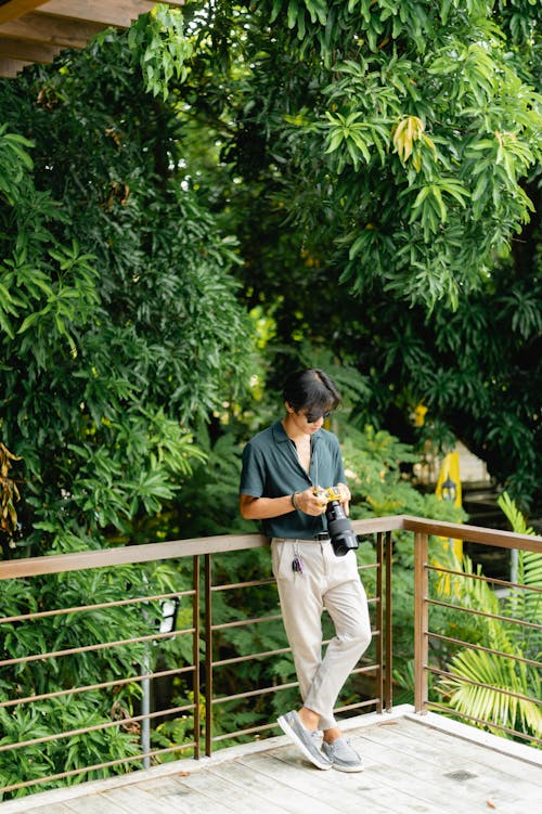 Young Man in Gray Short Sleeved Polo Standing and Leaning Against a Metal Railings Holding a Camera