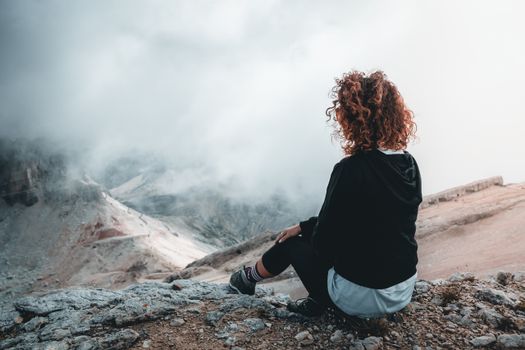 Woman sitting on rocky terrain, embracing the serene mountain and cloudscape view.