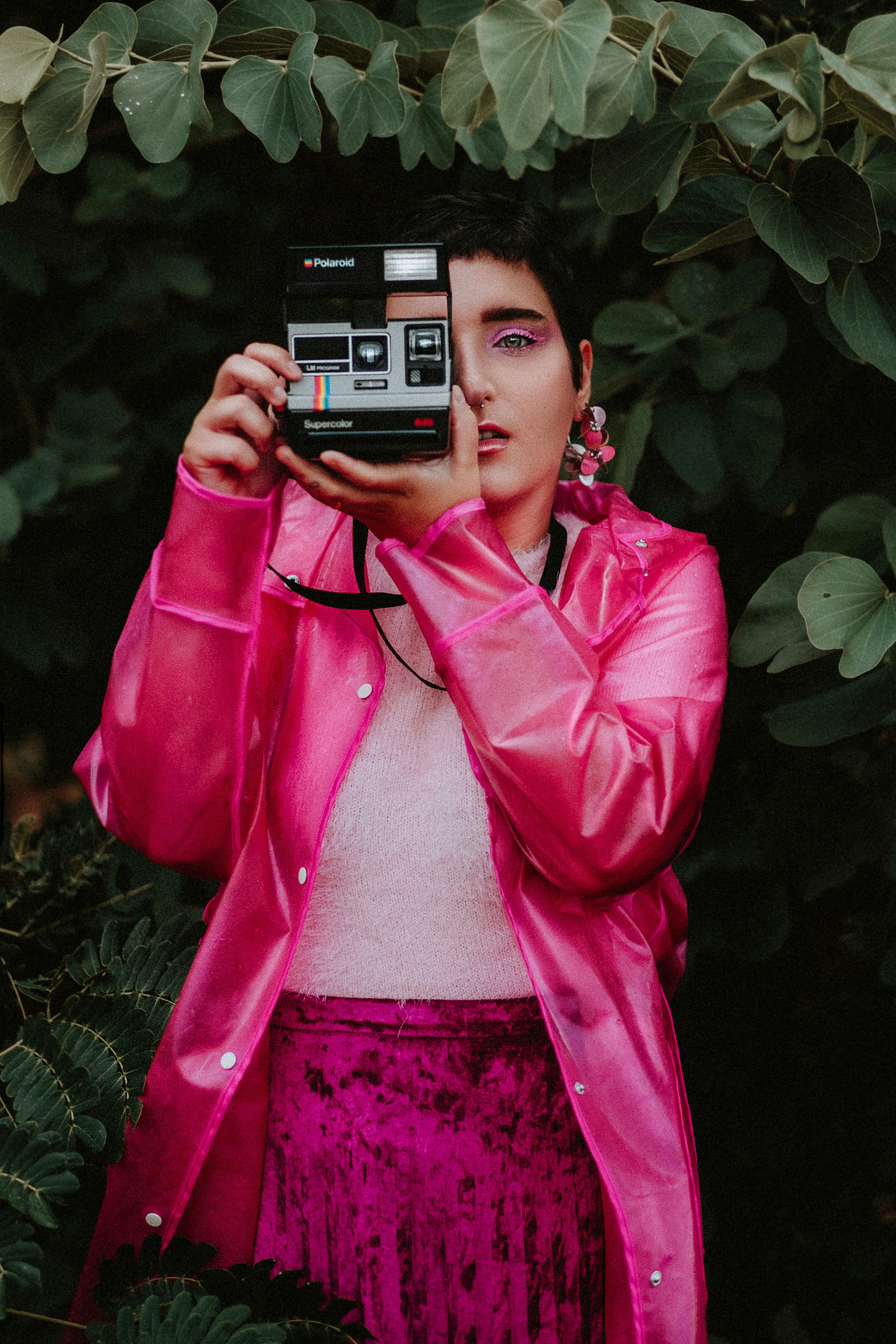 Stylish woman in pink coat using a polaroid camera among lush greenery.