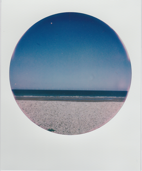 Circular polaroid capturing a tranquil beach and sea under a clear blue sky.