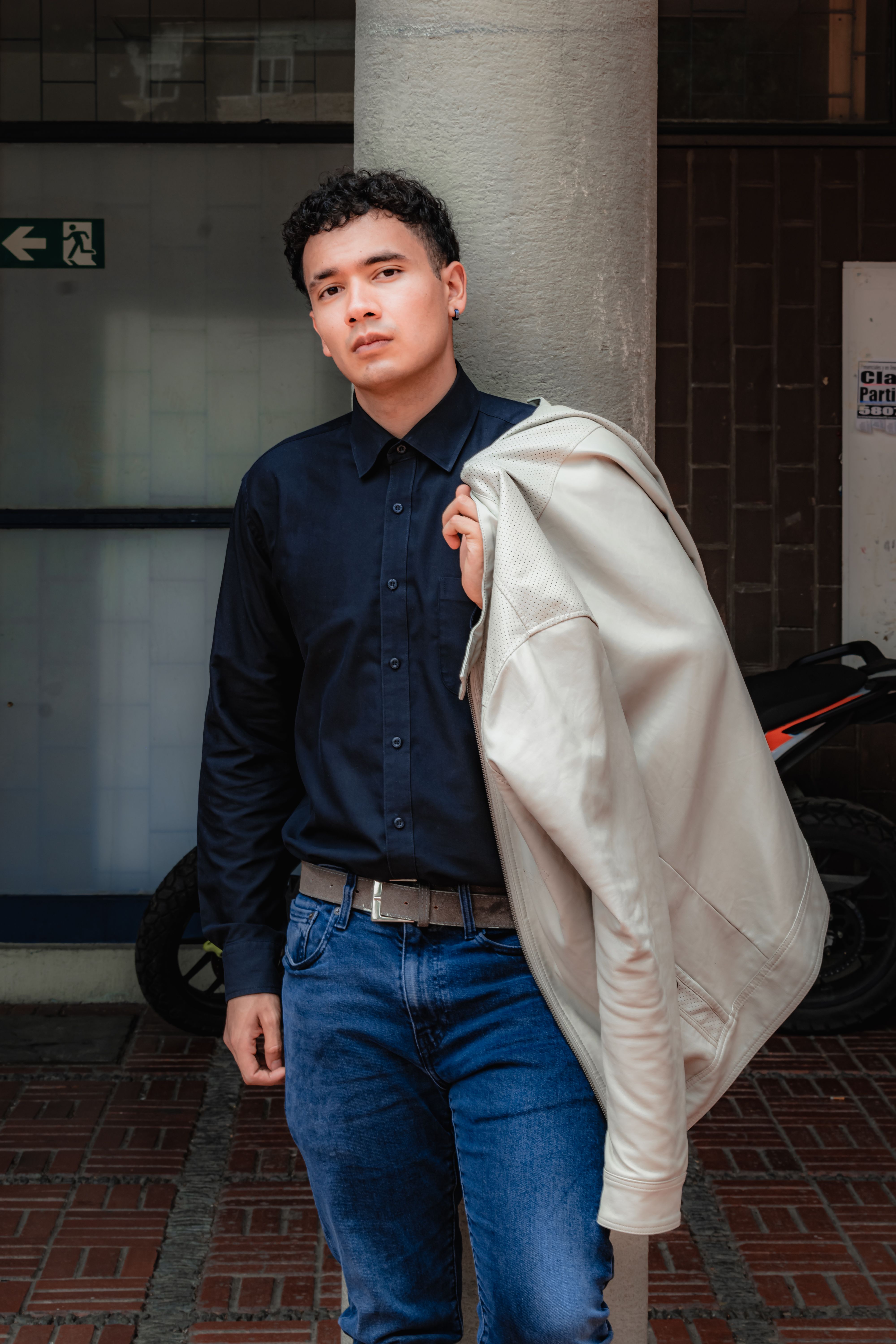 A young man poses confidently with a jacket slung over his shoulder in an urban environment.