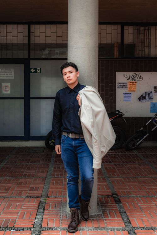 Free A casually dressed young man leans against a pillar, holding a jacket. Stock Photo