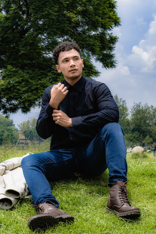 Free Young man sitting on grass in park wearing casual outfit, surrounded by nature. Stock Photo