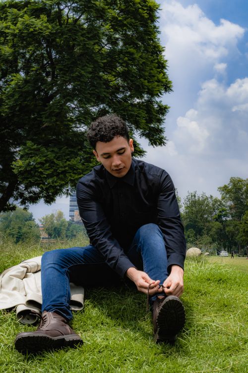 Free A young man sits on grass tying his boots in a sunny park with trees and blue sky. Stock Photo
