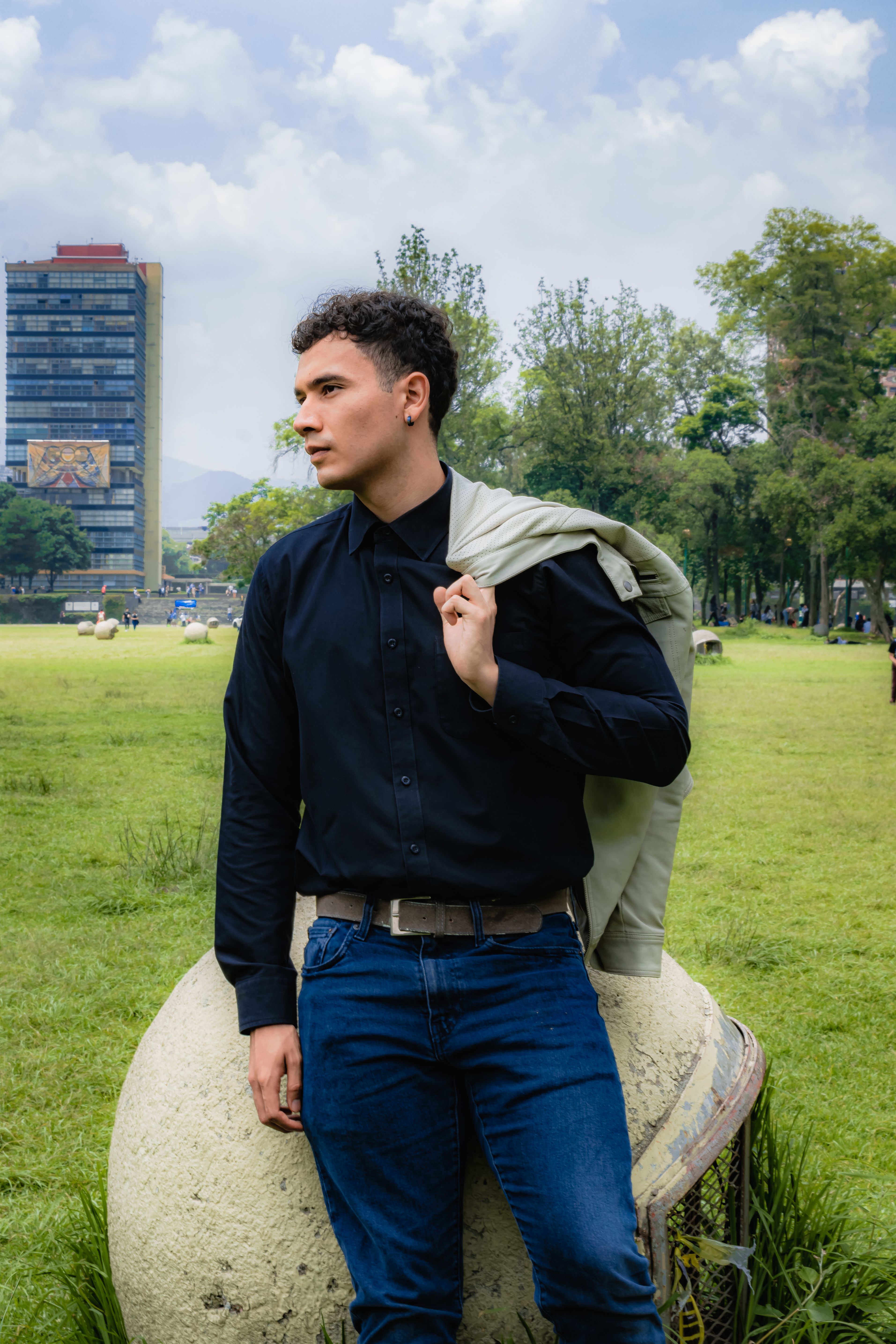 Man in casual attire poses confidently in a city park with a modern building in the background.