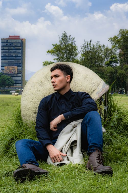 Free A fashionable young man in black attire sitting on green grass with a building in the background. Stock Photo